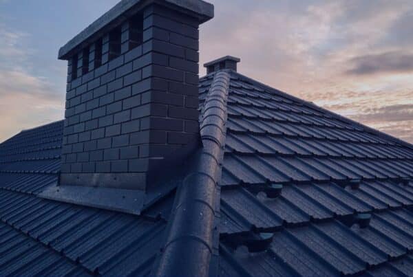 A high-angle shot of a modern dark metal tile roof featuring a large brick chimney and a ridged hip line against a sunset sky.
