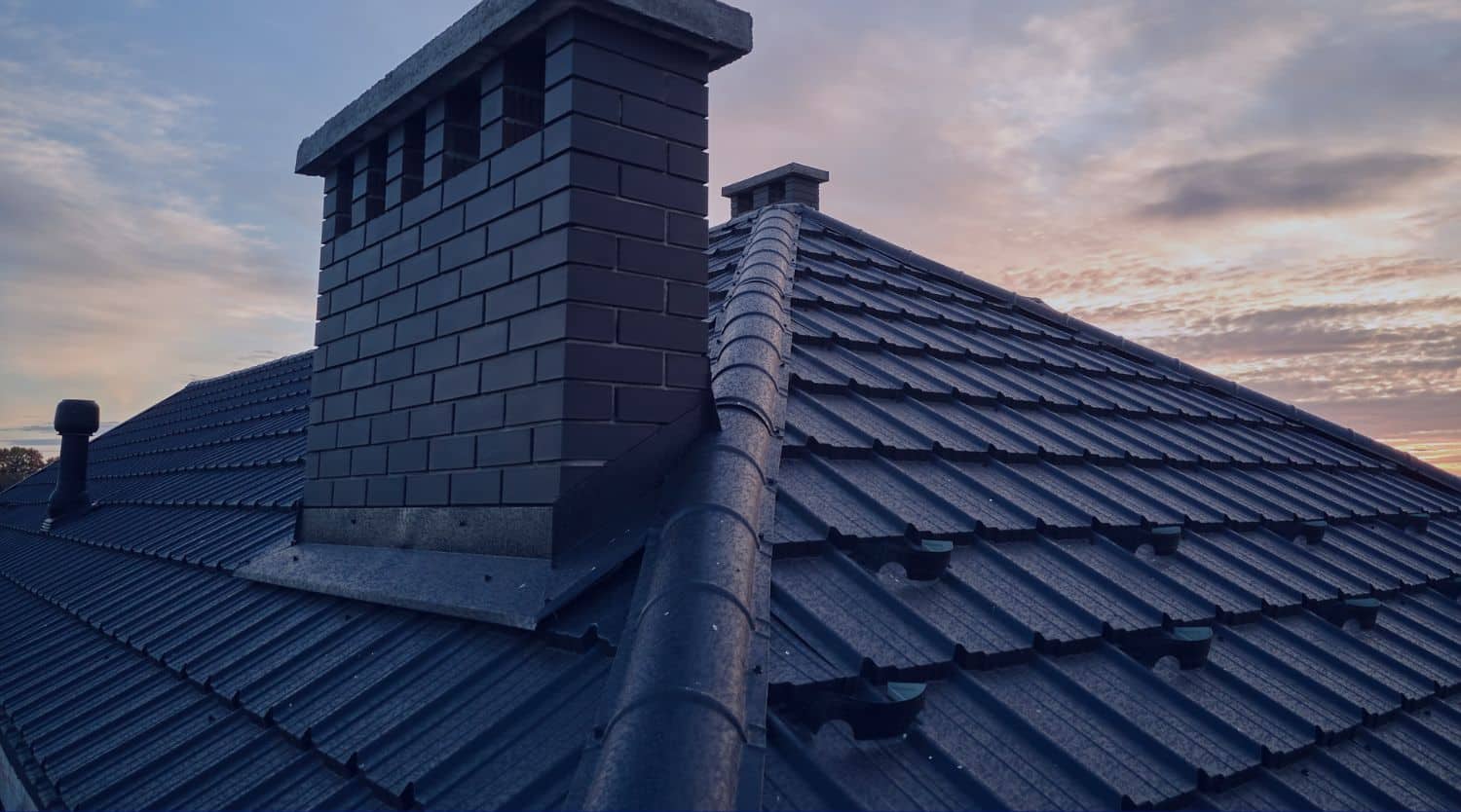 A high-angle shot of a modern dark metal tile roof featuring a large brick chimney and a ridged hip line against a sunset sky.