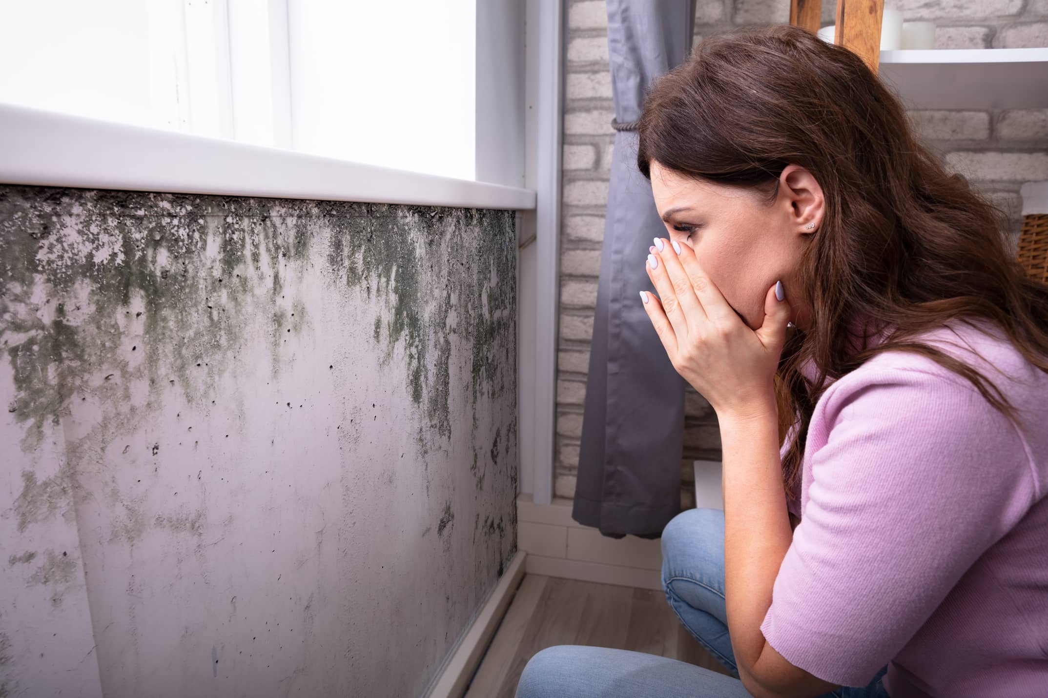 Concerned woman covering her mouth while inspecting black mold growth on a wall beneath a window.