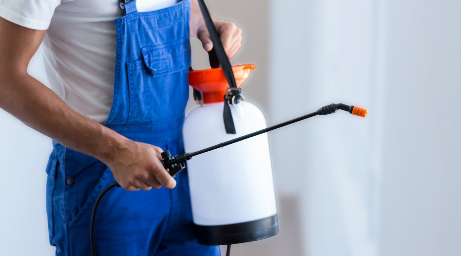 A person wearing blue overalls holding a white pump pressure sprayer tank and wand.